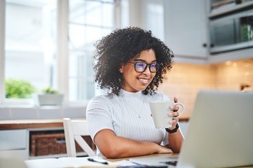 Eine Frau sitzt Zuhause an Ihrem Schreibtisch vor dem Laptop lächelnd mit einer Tasse in der Hand.