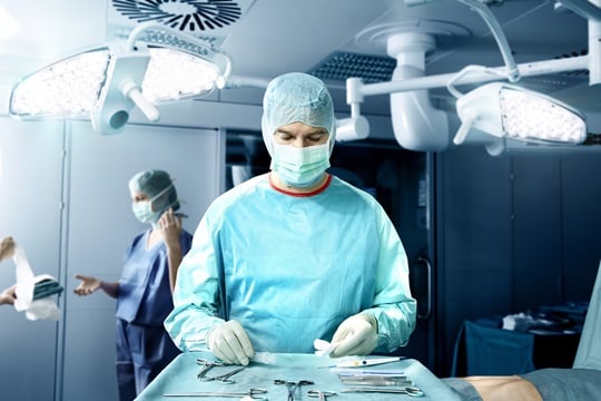 A doctor, wearing a surgical gown, is bent over a surgical table with operation instruments, while a doctors assistant is giving another assistant sterile gloves in a surgery room.