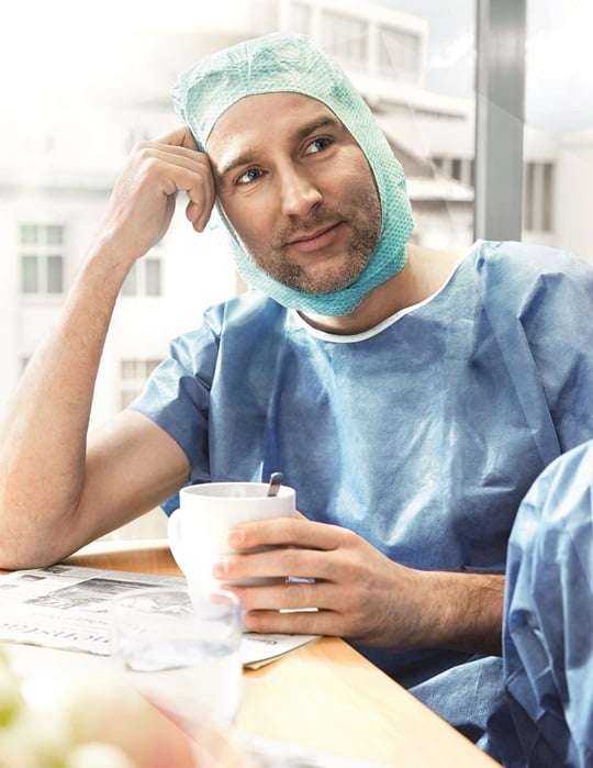 A doctor sits at a table and looking happy to a person in the Off. He put his head on his one hand and holding a cup in the other hand. He is still wearing his surgical gown, even he’s sitting in the cafeteria.